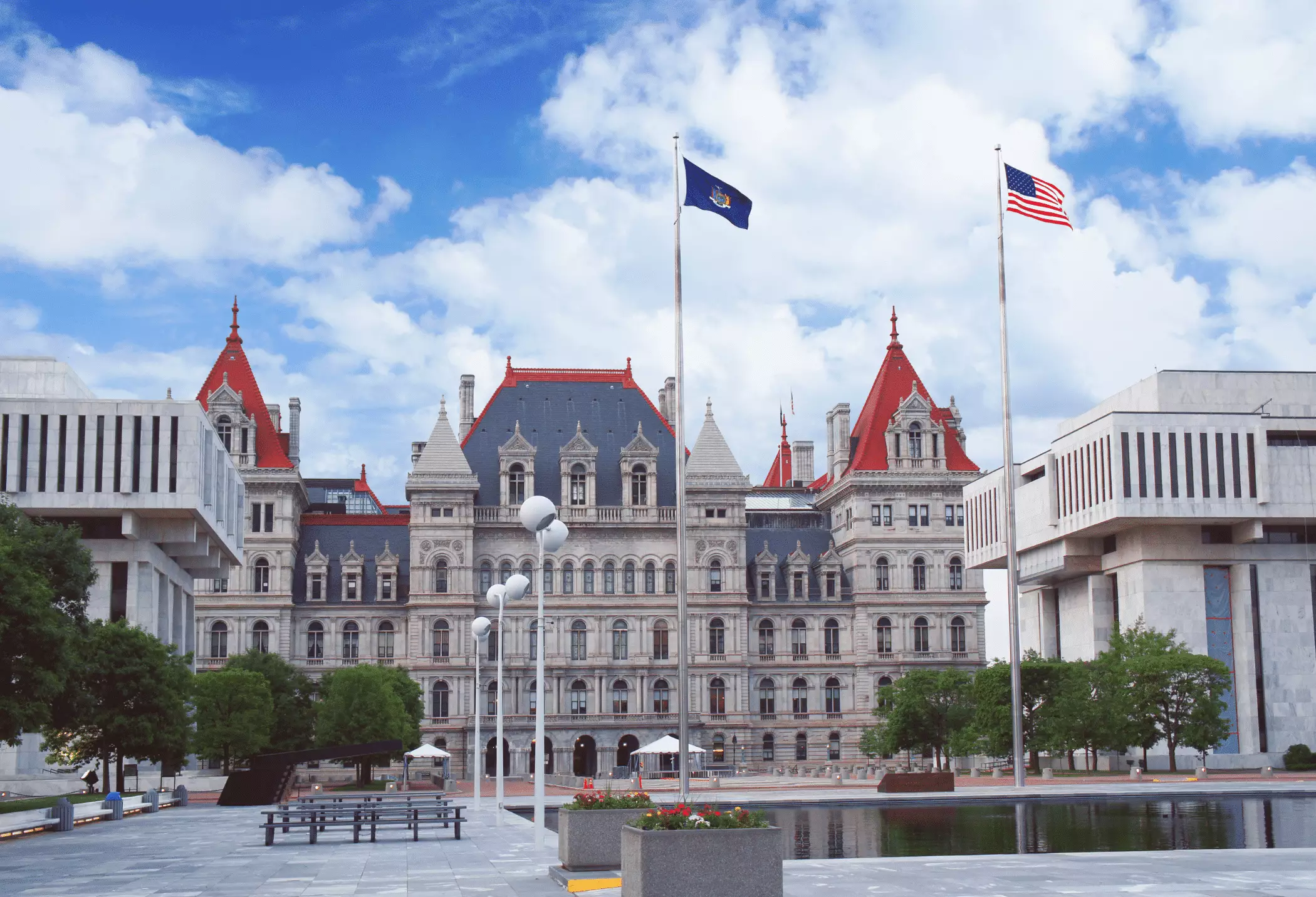 A grand historic government building with red roofs and stone masonry, flanked by modern architecture and a reflecting pool.