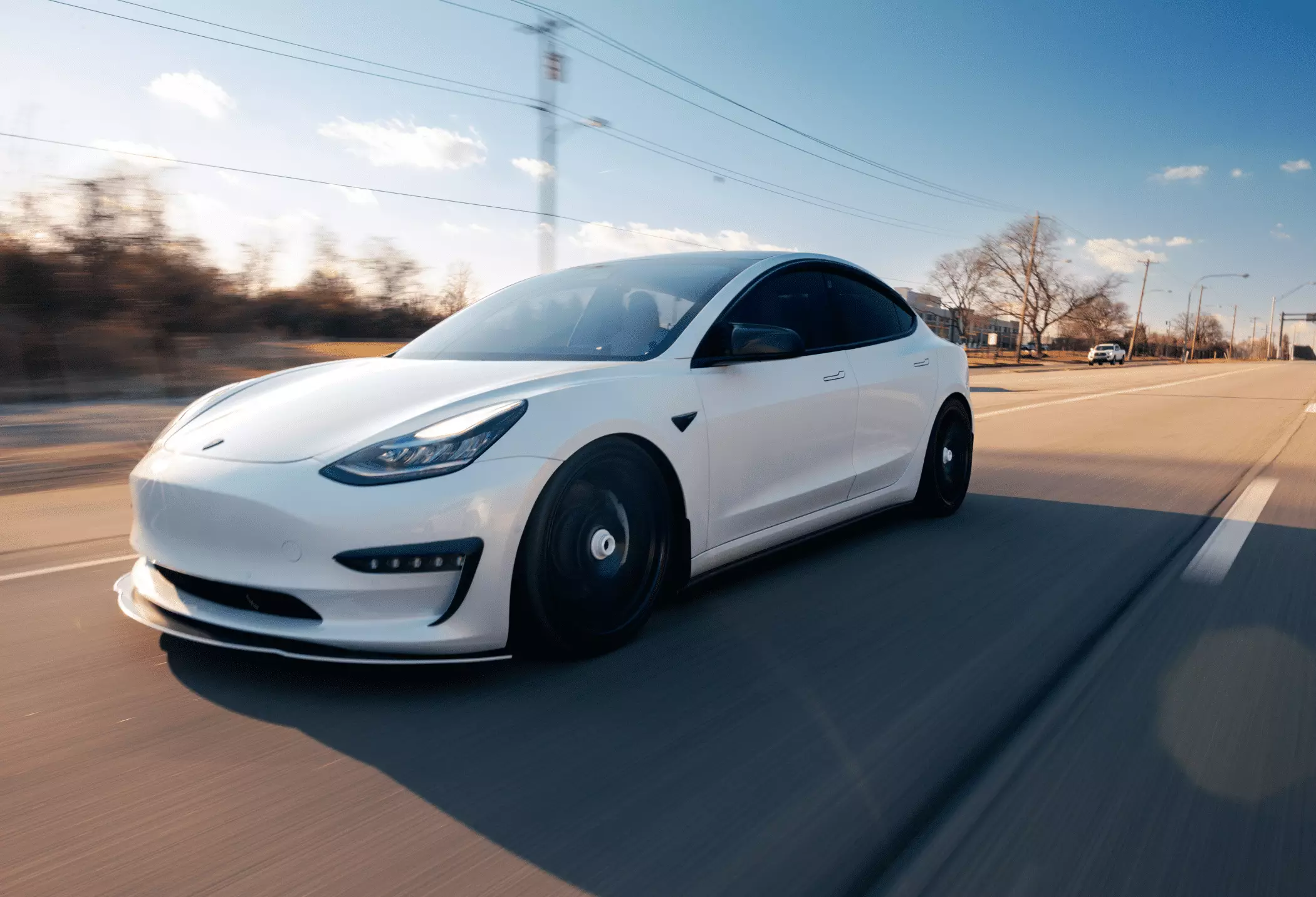 A white Tesla Model 3 driving quickly down a multi-lane road during the late afternoon.