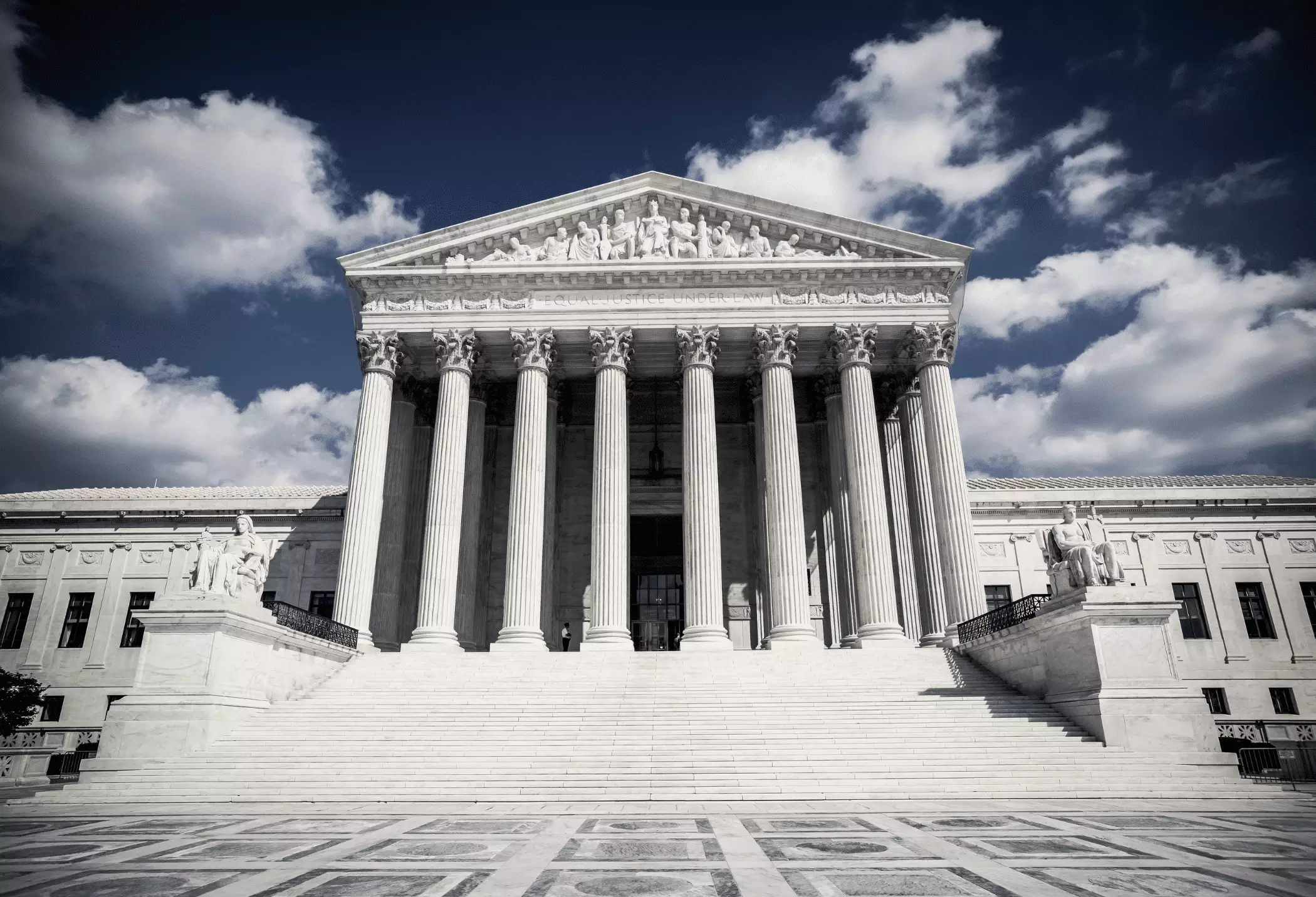 A wide, low-angle view of the United States Supreme Court building with tall Corinthian columns, stone statues, and a dramatic cloudy sky in the background.